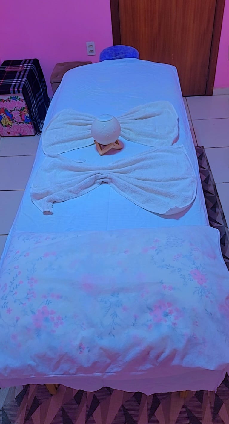 Detalhe da maca vista de cima, com lençol floral, toalhas brancas em laço e luminária de sal no centro